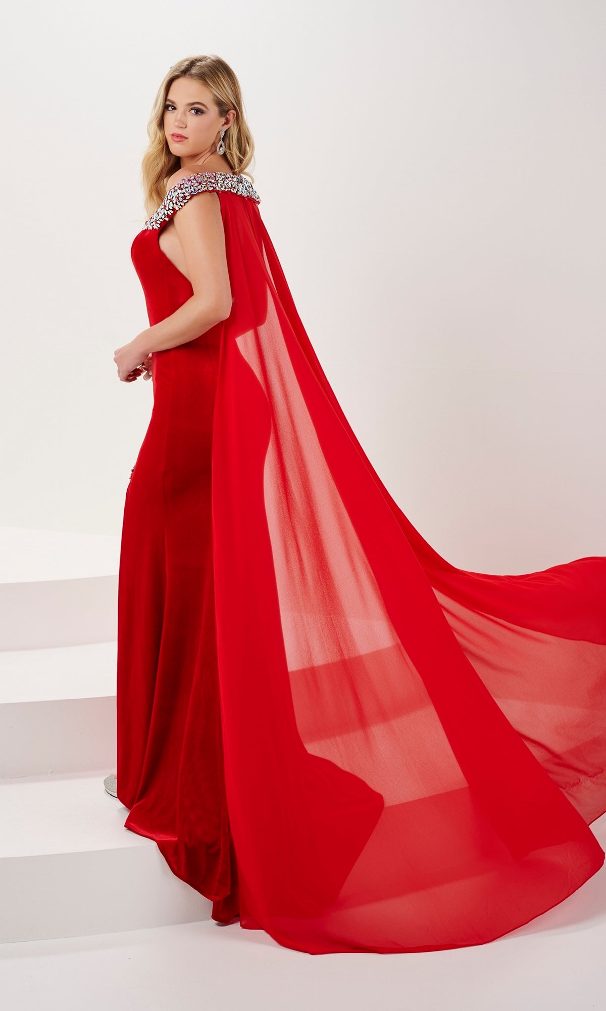 A woman in the Long Prom Dress 14195 by Panoply—a fitted red off-the-shoulder gown featuring a jeweled neckline and long sheer cape—stands on white steps, glancing over her shoulder.